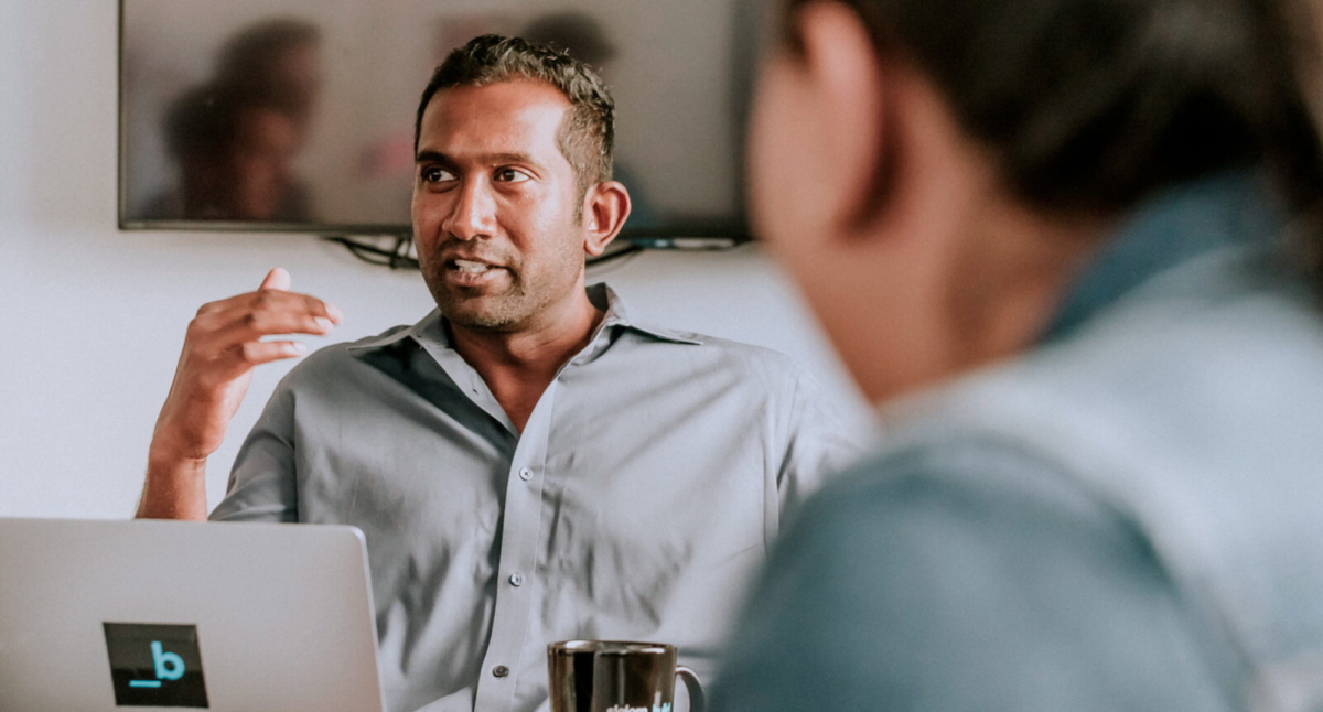 Man talking and gesturing at a conference table in front of a laptop.